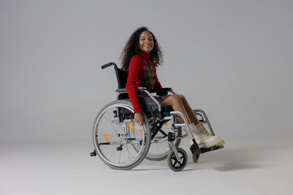 Young girl smiling in a wheelchair, wearing a plaid dress and boots against a white background.